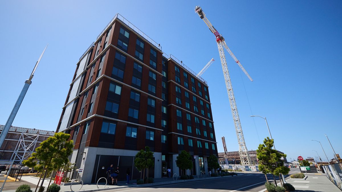 Low-angle view of a modern mid-rise building with dark windows and vertical brown paneling beside a tall construction crane under a clear blue sky; a quiet street with small trees, bike racks, and a f...