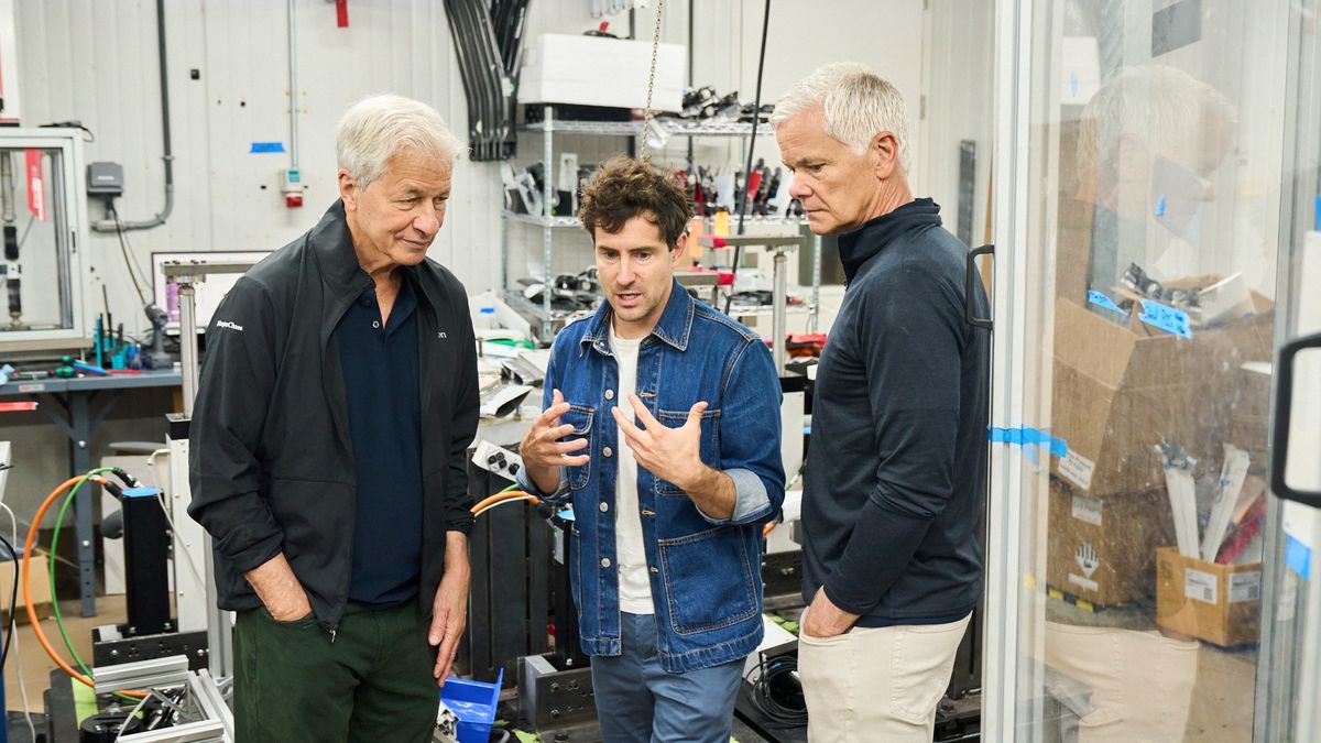 JPMorganChase CEO Jamie Dimon and two men talk in a workshop filled with equipment, tools, and parts shelves.