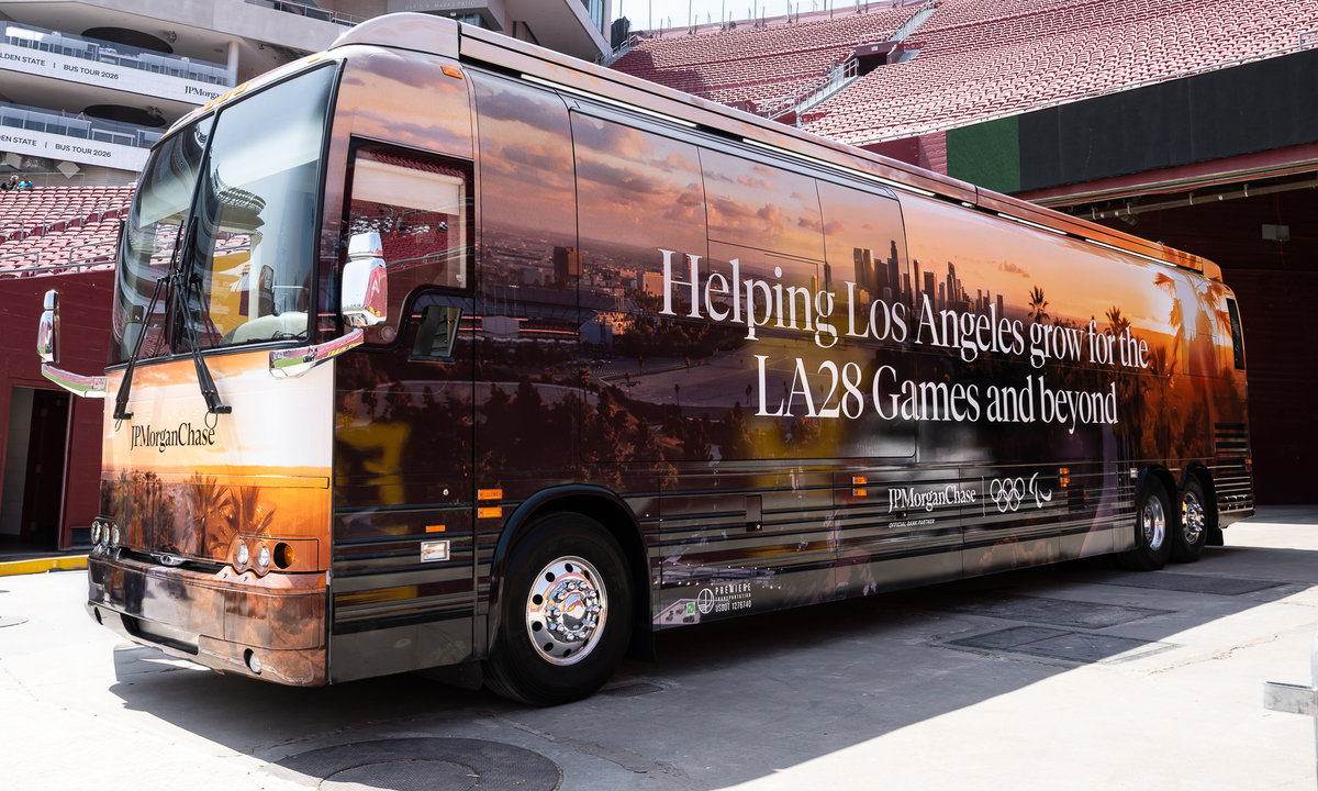 A large JPMorganChase coach bus parked inside a stadium concourse, wrapped with a sunset cityscape and the text "Helping Los Angeles grow for the LA28 Games and beyond."