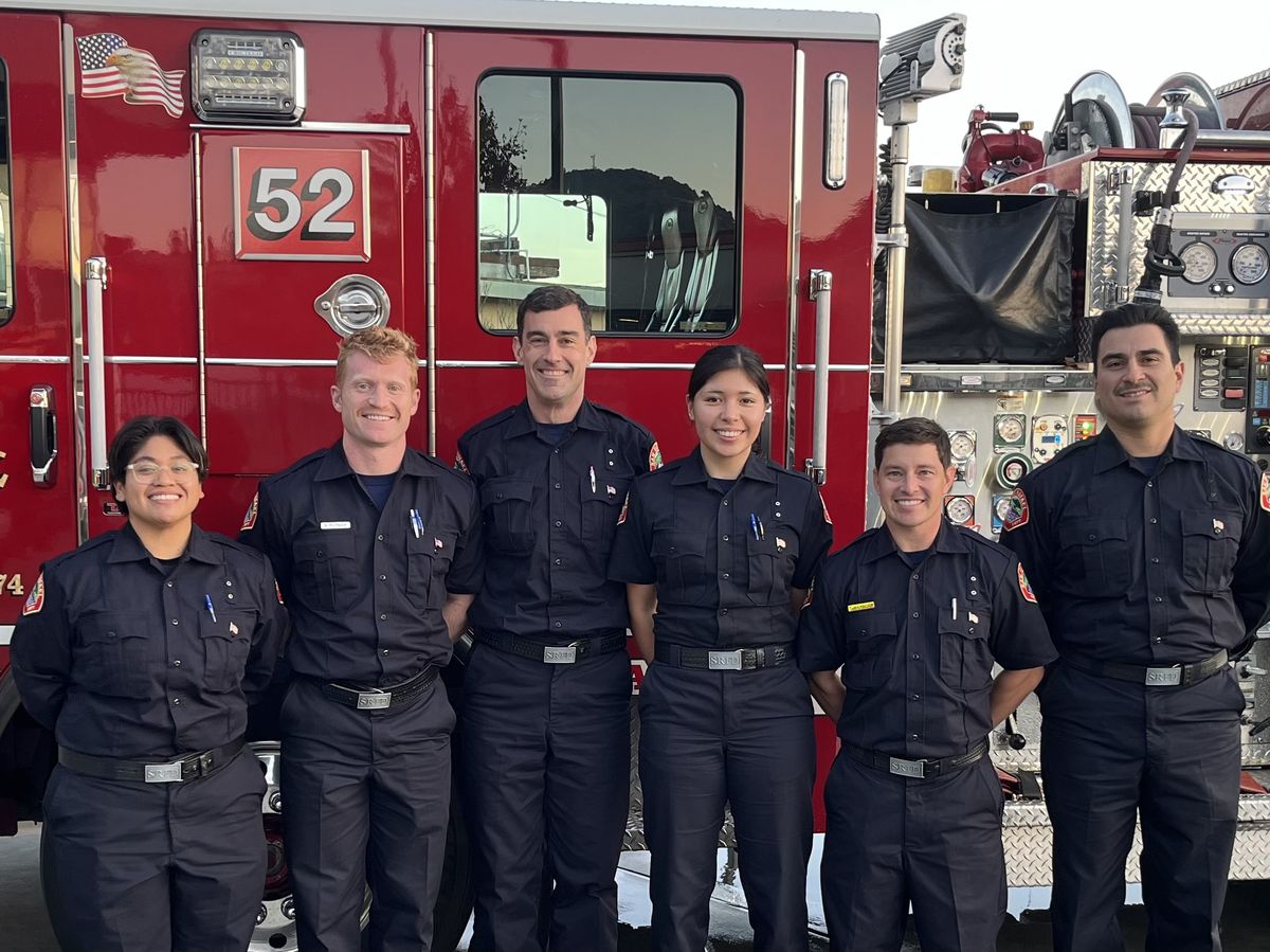 Photo of five smiling people in black uniforms in front of a fire engine