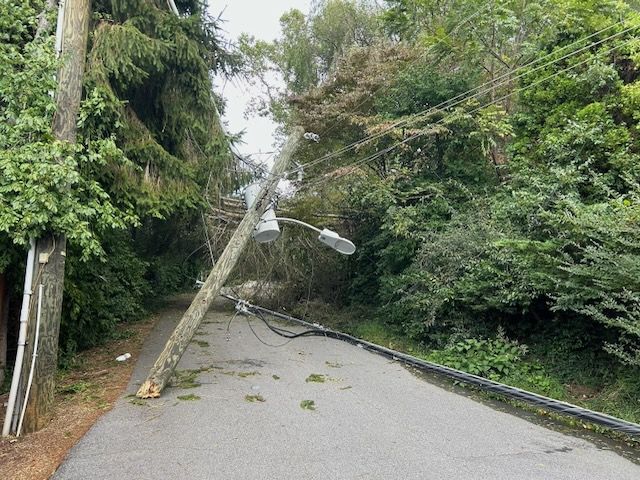 A utility pole with power and cable lines, a street light and a transformer still attached to it, broken in half and lying in a street in front of a fallen tree across the street