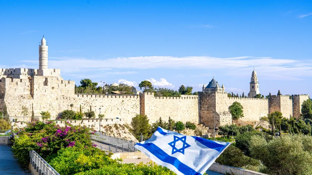 A panoramic view of the walls of the Old City of Jerusalem and the Tower of David, with the Israeli flag in the foreground.  Shutterstock