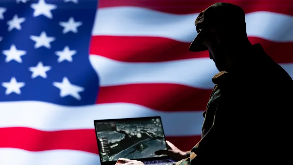 U.S. flag backdrop with uniformed soldier looking at laptop in foreground. Shutterstock