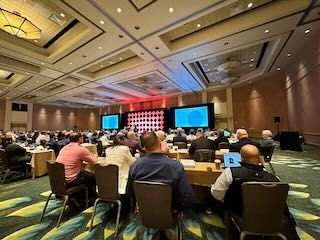 People sitting in a large conference room facing a stage and two large screens