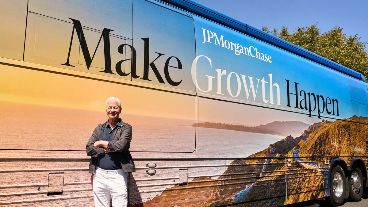 A man (Jamie Dimon, Chairman and CEO of JPMorganChase) stands with arms crossed in front of a large blue JPMorganChase bus wrapped with a coastal landscape and the slogan "Make Growth Happen."