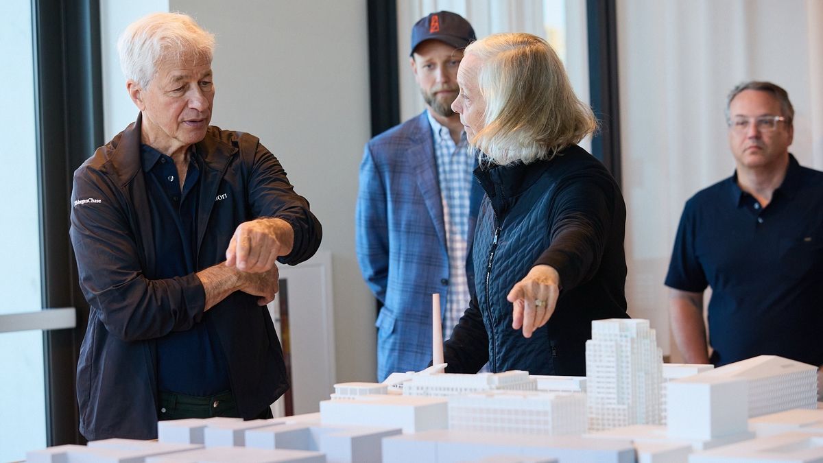 Two people stand over a white architectural scale model in a bright room, both gesturing and pointing as they discuss, while two others observe in the background.