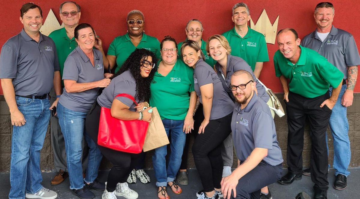 13 people in gray and green shirts in front of a red wall